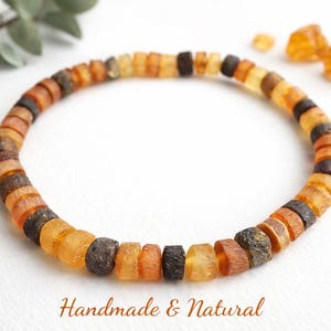 Peut inclure: Un bracelet en ambre fait à la main, composé de perles orange, marron et noires. Le bracelet est présenté sur une surface blanche avec une branche de verdure. Le texte "Handmade & Natural" est visible.