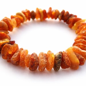May include: An amber bracelet. The bracelet is composed of irregular, polished amber stones in shades of orange and brown. The stones are strung together to form a circular shape. The bracelet is displayed against a white backdrop. The amber stones are of varying sizes.