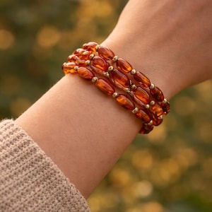 Op de afbeelding: Drie amberkleurige armbanden met ovale kralen, gescheiden door kleine goudkleurige kralen. De armbanden worden om een pols gedragen. De achtergrond is wazig, met groene en gele tinten. De kralen zijn ongeveer 1 cm lang.