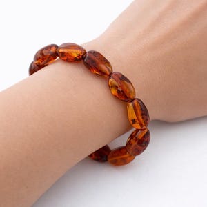 Könnte beinhalten: Ein Armband aus polierten Bernsteinperlen, jede oval geformt und in verschiedenen Orange- und Brauntönen gehalten. Das Armband wird am Handgelenk getragen und zeigt die natürliche Farbe und Textur des Bernsteins. Die Perlen sind etwa 4 cm lang.