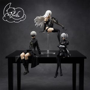 May include: Three detailed figurines of characters from the Nier: Automata video game. The figures are in black and white, with one figure seated on a clear stand. The figures are posed on a black table against a dark grey background.