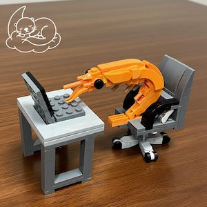 Puede incluir: Una escena Lego caprichosa presenta una figura de camarón naranja sentada en una silla de oficina gris, trabajando en una pequeña computadora portátil gris en un escritorio blanco. Un contorno blanco de una nutria durmiendo en una nube está en el fondo. La escena está sobre una superficie marrón.