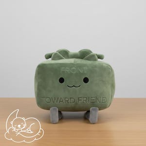 May include: A green plush toy with the words "FRONT" and "TOWARD FRIEND" embroidered on it. The toy has a smiling face with two black eyes and a curved mouth. It has two small, grey legs and two green protrusions on top.