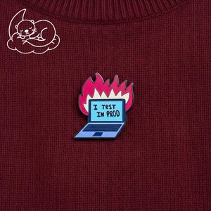May include: A burgundy jumper with a white cartoon otter graphic and a laptop pin. The pin features a blue laptop with the text "I TEST IN PROD" surrounded by pink flames. The jumper has a ribbed texture.