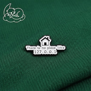 May include: A black and white enamel pin with the text "There is no place like 127.0.0.1" and a house graphic. The pin is on a dark green knitted fabric. A white cartoon otter is in the upper left corner.