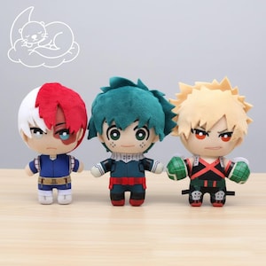 May include: Three plush dolls of anime characters. The dolls have large eyes and detailed outfits. One doll has red and white hair, another has green hair, and the third has spiky blonde hair. The dolls are standing on a light brown surface.