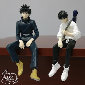 Puede incluir: Dos figuras de estilo anime de jóvenes sentados con las piernas colgando. Uno viste un atuendo azul oscuro con botas amarillas, el otro una camisa blanca, pantalones negros y zapatillas blancas. Ambos tienen cabello negro y están posados sobre una superficie marrón.