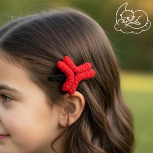 May include: A red, crocheted hair clip in the shape of a star is attached to a person's hair. The hair clip is made of a textured yarn and is secured with a black metal clip. The background is blurred, with a green and brown color scheme.
