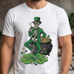 Op de afbeelding: Wit t-shirt met een skelet in een groen pak en hoed, een pijp rokend, zittend op een pot goud. Het ontwerp bevat een klavertjevier en gouden munten, perfect voor St. Patrick's Day.