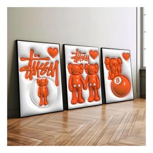 May include: Three framed art pieces featuring orange 3D designs on white backgrounds. The art includes the "Stussy" logo, heart shapes, and cartoon figures. The frames are black, and the artwork is displayed on a wooden floor.