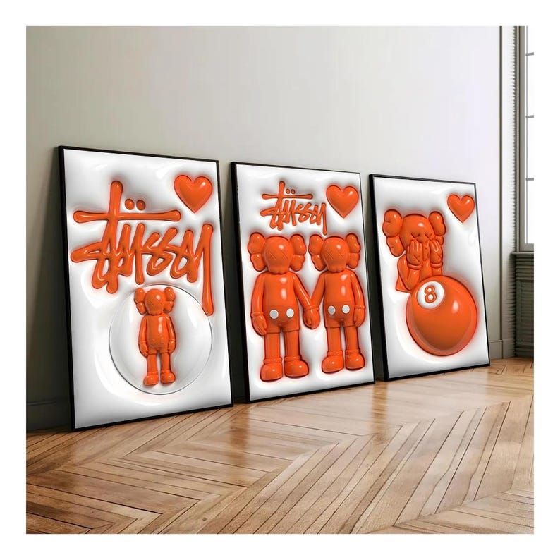 May include: Three framed art pieces featuring orange 3D designs on white backgrounds. The art includes the "Stussy" logo, heart shapes, and cartoon figures. The frames are black, and the artwork is displayed on a wooden floor.