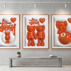 May include: Three framed art pieces with orange accents. The artwork features the Stussy logo, KAWS figures, and a heart. The frames are orange, and the background is white. The art is displayed above a gray bench.