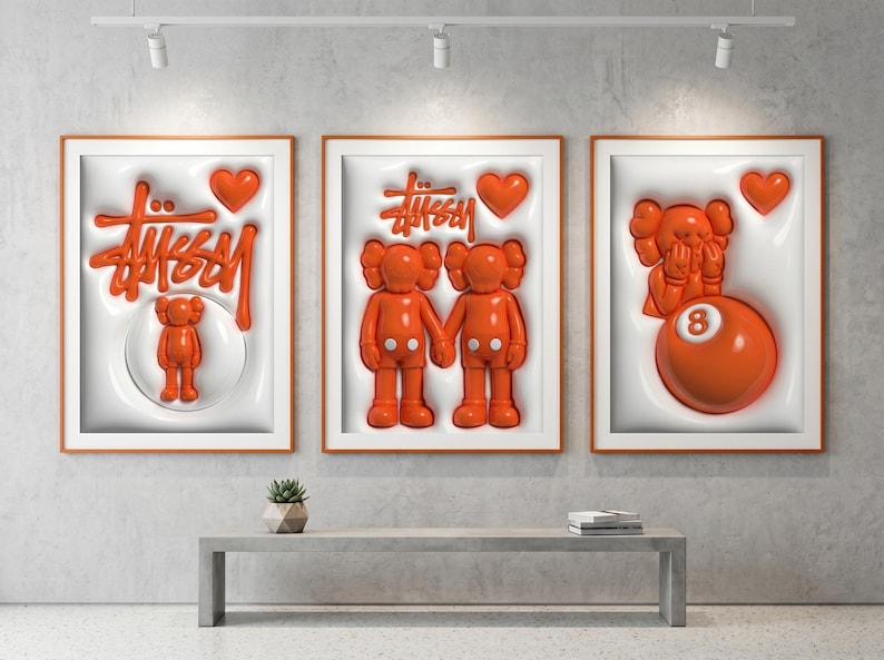 May include: Three framed art pieces with orange accents. The artwork features the Stussy logo, KAWS figures, and a heart. The frames are orange, and the background is white. The art is displayed above a gray bench.