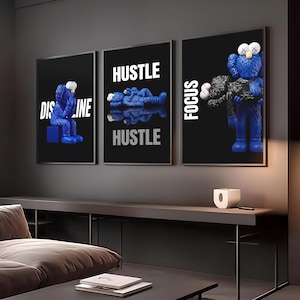 May include: Three framed art prints with black backgrounds. Each features a blue cartoon figure. One print says "DISCIPLINE", another says "HUSTLE" twice, and the third says "FOCUS". The art is displayed above a dark gray console table.