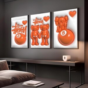 May include: Three framed wall art pieces featuring orange and white designs. The art includes the Stussy logo, KAWS figures, hearts, and a billiard ball with the number 3 and 8. The artwork is displayed on a dark gray wall.