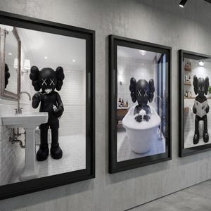 May include: Three framed art pieces featuring a black figure in a bathroom setting. The figure is depicted brushing its teeth, bathing in a tub, and reading a newspaper. The frames are black and mounted on a gray wall.