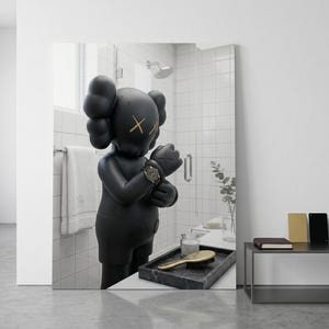 May include: A matte black KAWS figure with signature X-eyes and gloved hands stands in a tiled bathroom. The figure wears a dark wristwatch on its left wrist. A black marble tray with a gold-toned hairbrush and a glass jar sits in the foreground.