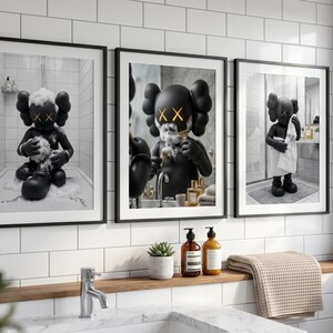 May include: A set of three framed wall art prints featuring a black KAWS character in a bathroom setting. The first shows the figure bathing, the second shaving in a mirror, and the third drying off with a white towel. Each print has a black frame.