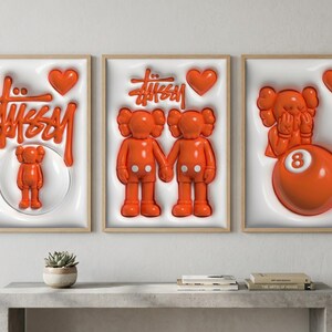 May include: Three framed art prints with orange and white elements. The prints feature the Stussy logo, Kaws figures, and an 8-ball. Each print has a wooden frame and a white background with a 3D effect. The art pieces are displayed above a gray table.