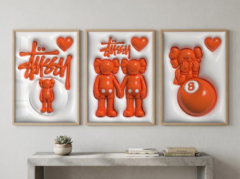 May include: Three framed art prints with orange and white elements. The prints feature the Stussy logo, Kaws figures, and an 8-ball. Each print has a wooden frame and a white background with a 3D effect. The art pieces are displayed above a gray table.