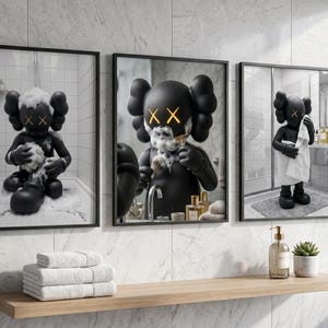 May include: A set of three framed wall art prints featuring a black cartoon character with X-shaped eyes in a bathroom setting. The prints show the character bathing, shaving, and drying off with a white towel, all rendered in a monochromatic style.