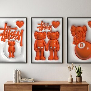 May include: Three framed art prints featuring orange pop art designs. One print displays the Stussy logo and a small figure, another shows two figures holding hands, and the third has a figure and an orange 8-ball.