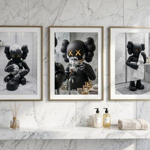 May include: A set of three framed wall art prints featuring a black cartoon character with X-shaped eyes in a bathroom setting. The first shows the character bathing, the second shaving, and the third drying off with a white towel in a marble room.