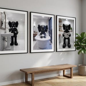 May include: Three framed art prints featuring a black figure with crossed-out eyes in bathroom settings. One print shows the figure at a sink, another in a bathtub, and the third on a toilet reading a newspaper. The prints are displayed above a wooden bench.