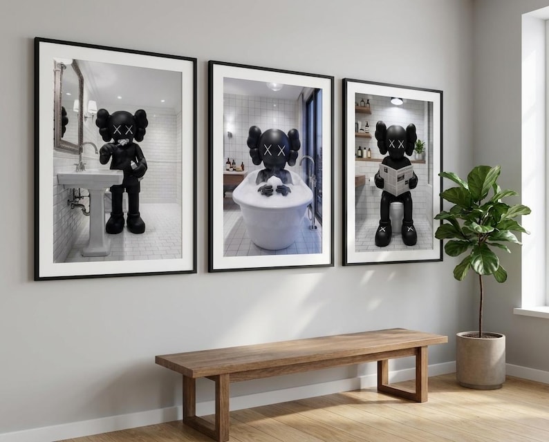 May include: Three framed art prints featuring a black figure with crossed-out eyes in bathroom settings. One print shows the figure at a sink, another in a bathtub, and the third on a toilet reading a newspaper. The prints are displayed above a wooden bench.