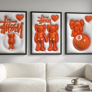 May include: Three framed art pieces with black frames. Each features a white background with orange elements, including the Stussy logo, hearts, and cartoon figures. A sofa and coffee table are in the foreground.