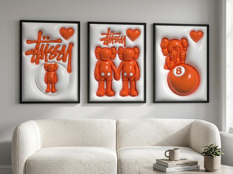 May include: Three framed art pieces with black frames. Each features a white background with orange elements, including the Stussy logo, hearts, and cartoon figures. A sofa and coffee table are in the foreground.