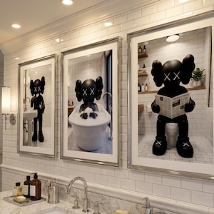 May include: Three framed art prints featuring a black figure with crossed-out eyes in bathroom settings. One print shows the figure standing at a sink, another in a bathtub, and the third on a toilet reading a newspaper. The prints are displayed above a marble countertop.