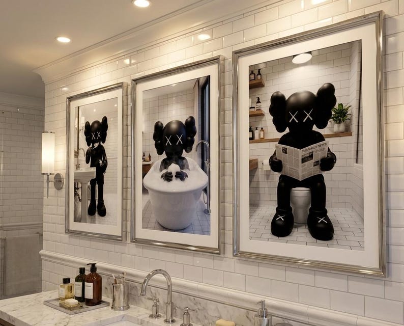 May include: Three framed art prints featuring a black figure with crossed-out eyes in bathroom settings. One print shows the figure standing at a sink, another in a bathtub, and the third on a toilet reading a newspaper. The prints are displayed above a marble countertop.