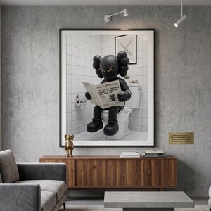 May include: A framed wall art print featuring a black KAWS character sitting on a white toilet while reading a newspaper titled The KAWS Times. The character has signature X-shaped eyes and rounded ears, set against a white tiled bathroom background.