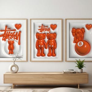 May include: Three framed art pieces with orange 3D designs. The art features the Stussy logo, KAWS figures, and an 8-ball. Each piece has a white background and a wooden frame. The art is displayed above a wooden cabinet.