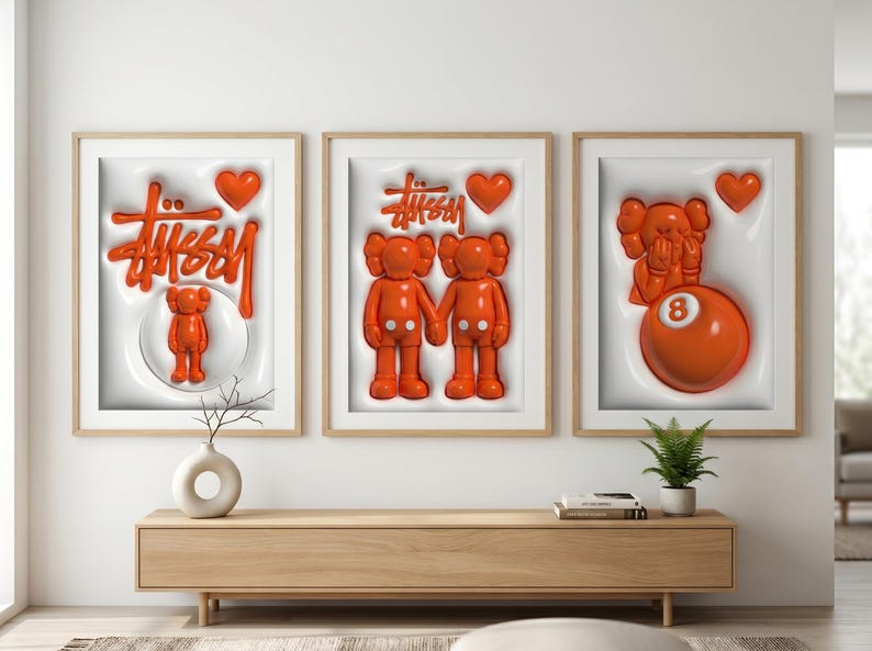 May include: Three framed art pieces with orange 3D designs. The art features the Stussy logo, KAWS figures, and an 8-ball. Each piece has a white background and a wooden frame. The art is displayed above a wooden cabinet.