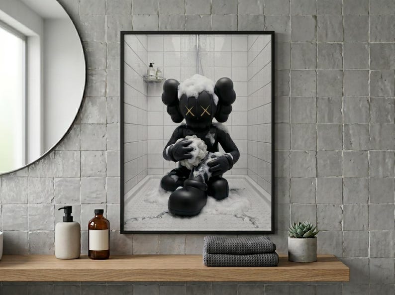 May include: A framed wall art print featuring a black cartoon character with X-shaped eyes sitting in a white tiled shower. The character is covered in white soap suds and is holding a sponge while water falls from a shower head above in the scene.