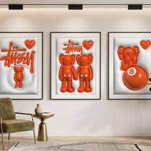 May include: Three framed art pieces with white backgrounds and black frames. Each features orange 3D designs, including the Stussy logo, hearts, and cartoon figures. One includes an orange 8-ball. The art is displayed above a green chair and side table.