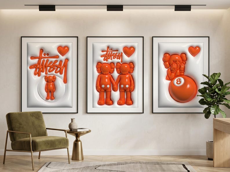 May include: Three framed art pieces with white backgrounds and black frames. Each features orange 3D designs, including the Stussy logo, hearts, and cartoon figures. One includes an orange 8-ball. The art is displayed above a green chair and side table.