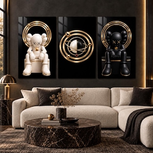 Op de afbeelding: Een set van drie wandkunstprints op een zwarte achtergrond. De linker print toont een wit cartoonfiguur met X-ogen zittend onder gouden ringen. De middelste print toont een zwart-witte bol met gouden orbitale ringen. De rechter print toont een zwart cartoonfiguur met X-ogen zittend onder gouden ringen.