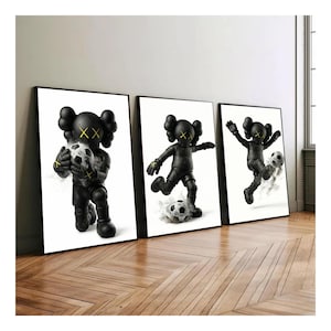 Op de afbeelding: Set van drie ingelijste kunstprints met een zwarte figuur met gele kruisogen en een voetbal. De figuur staat in verschillende actieposities tegen een witte achtergrond. De lijsten zijn zwart.