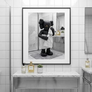 May include: A framed wall art print featuring a black KAWS character figure standing in a bathroom, wrapped in a white towel and holding another white towel to its face. The figure has yellow X symbols for eyes and is set against a white tiled wall.