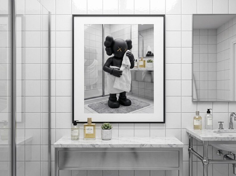May include: A framed wall art print featuring a black KAWS character figure standing in a bathroom, wrapped in a white towel and holding another white towel to its face. The figure has yellow X symbols for eyes and is set against a white tiled wall.