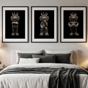 Pu&ograve; includere: Tre stampe artistiche incorniciate con una figura grigio scuro con una corona d'oro su sfondo nero. La figura &egrave; in diverse pose in ogni stampa. Le stampe sono esposte sopra un letto con lenzuola bianche e cuscini grigi.