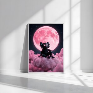 May include: A framed art print showing a black figure with X-shaped eyes and a black body, seated on a pink cloud. A large pink moon dominates the background, set against a starry night sky. The artwork is displayed in a white frame.
