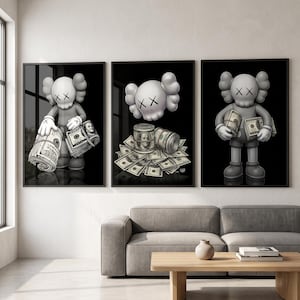 Op de afbeelding: Een set van drie ingelijste muurprints met een grijze stripfiguur met X-vormige ogen. De figuur houdt bundels opgerolde en platte Amerikaanse honderd-dollarbiljetten vast en is erdoor omringd, tegen een zwarte achtergrond.