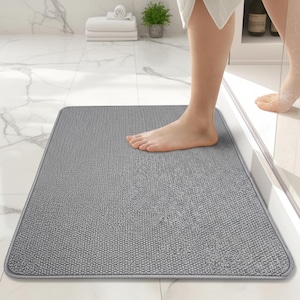 May include: A rectangular gray bath mat with a textured surface. The mat is placed on a white tile floor in a bathroom setting. A person's foot is on the mat, and another foot is visible near a shower.