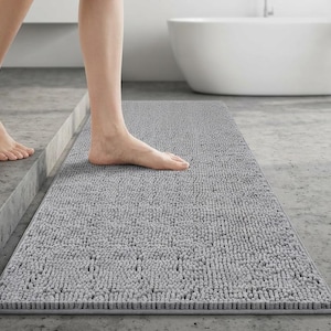 May include: A gray rectangular bath mat with a textured surface. The mat is placed on a concrete floor in a bathroom setting. A person is stepping onto the mat.
