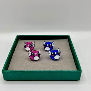 May include: Four small, round, cartoon-like pins with white, pink, and blue designs are displayed in a green box. The pins have a black outline and a white face with two black dots. The box has a gray interior.