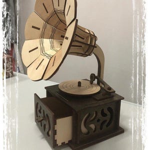 May include: A wooden model of a vintage gramophone. The horn is light brown, and the base is dark brown with an open drawer. The turntable and arm are also visible. The model is made of layered wood pieces.